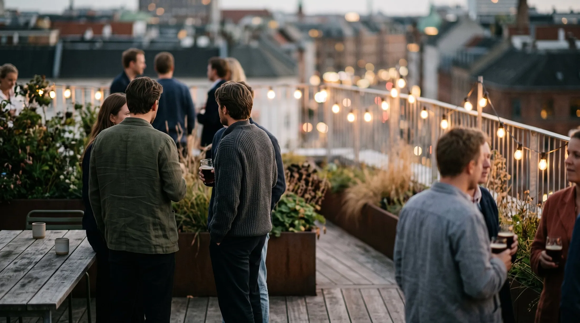 People in small groups on a rooftop terrace at dusk, string lights and rooftops behind