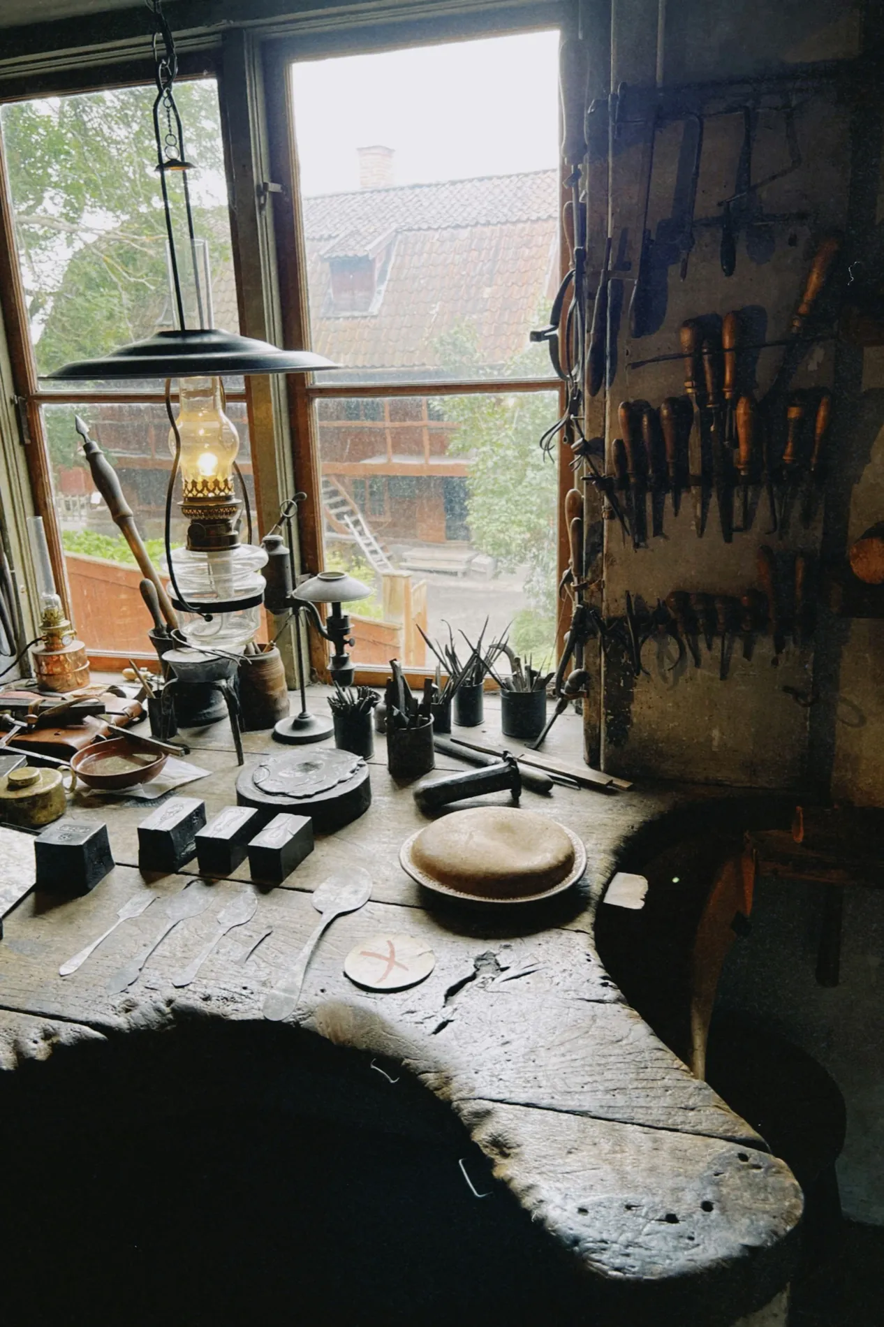 Een werkbank onder een raam, met een olielamp en handgereedschap over het versleten blad