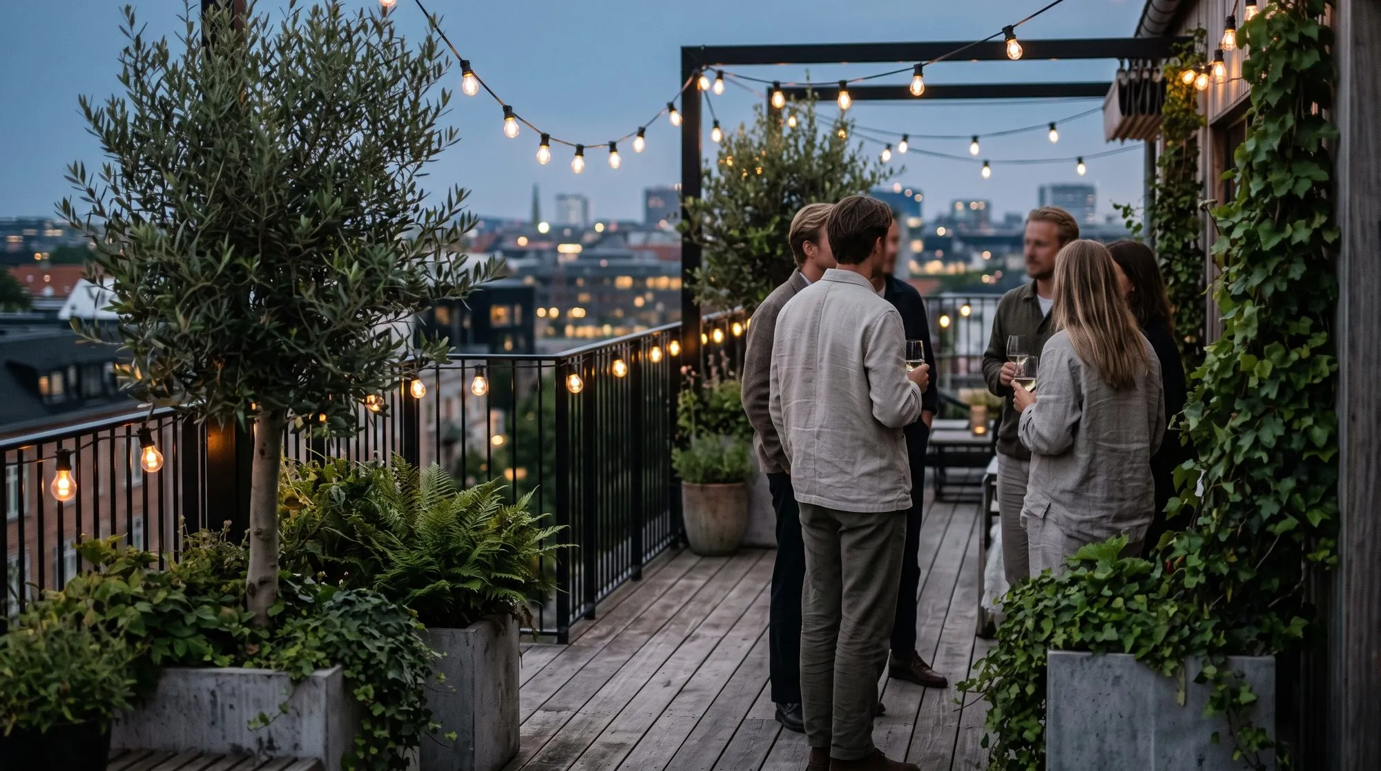 Two small groups talking on a rooftop terrace under festoon lights, an olive tree in a planter