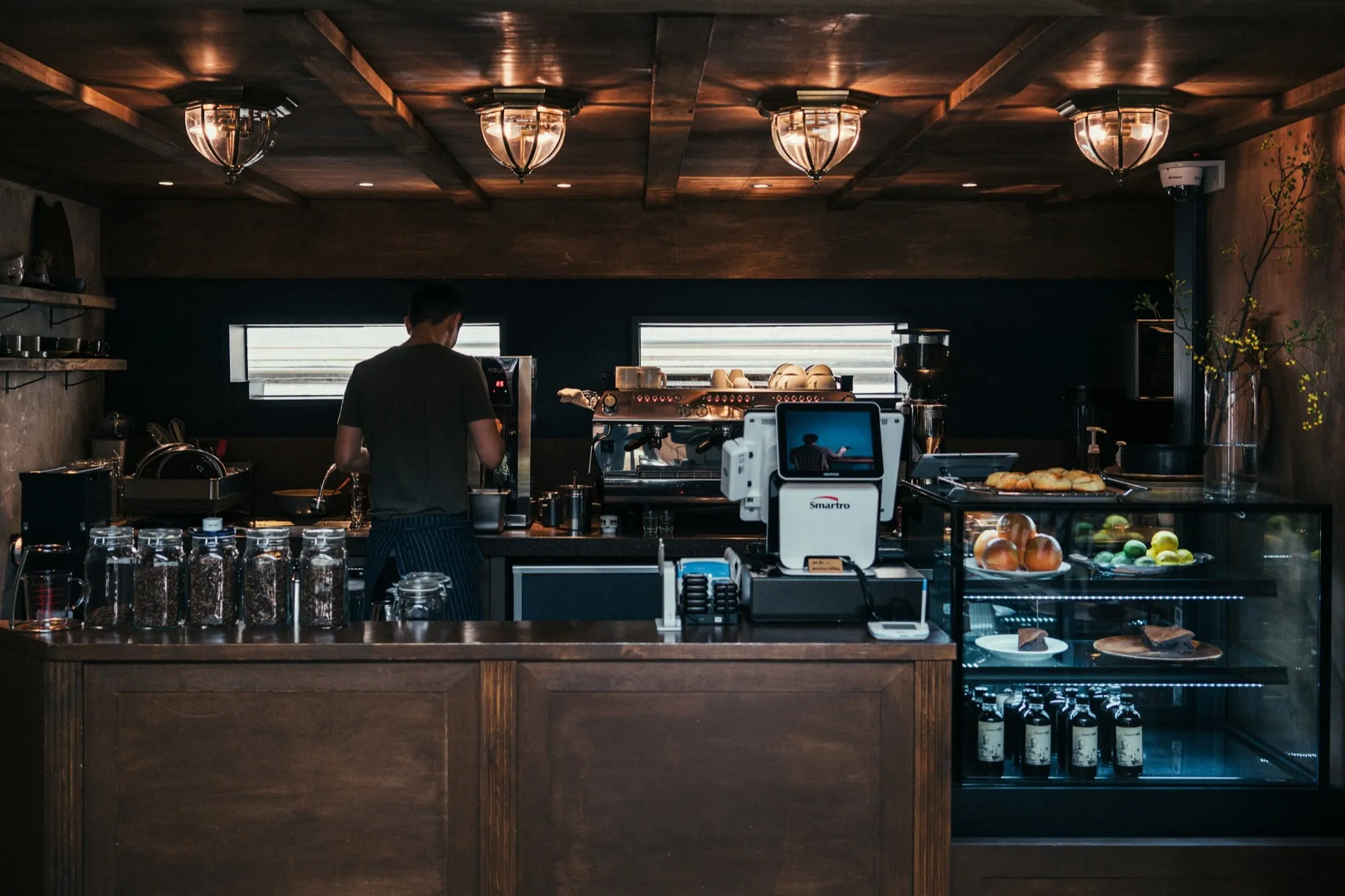 A cafe counter seen from the room: one person working behind the machine, warm lamps overhead