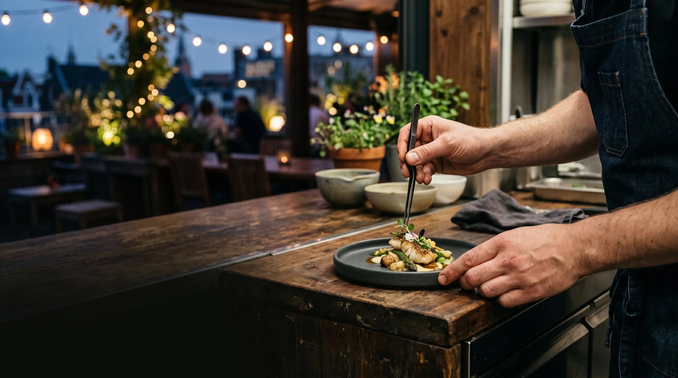 A cook finishing a plate on a rooftop kitchen at dusk, string lights and a canal behind