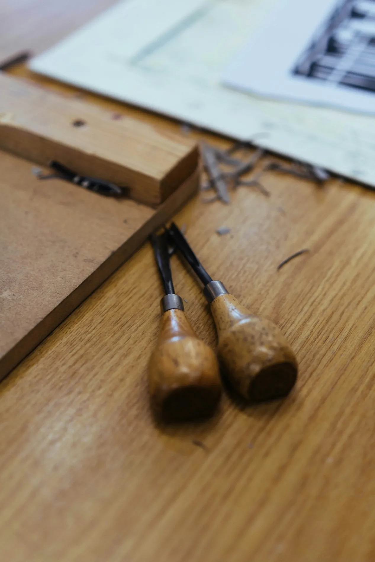 Two worn wooden-handled chisels lying on a bench beside a cut sheet and drawings
