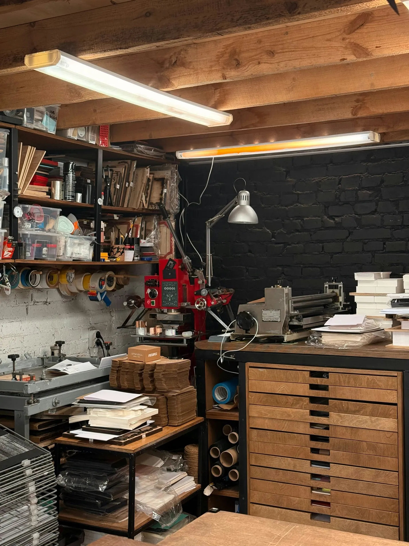 A print workshop: a plan chest, a press, inked rollers and stacked paper under a strip light