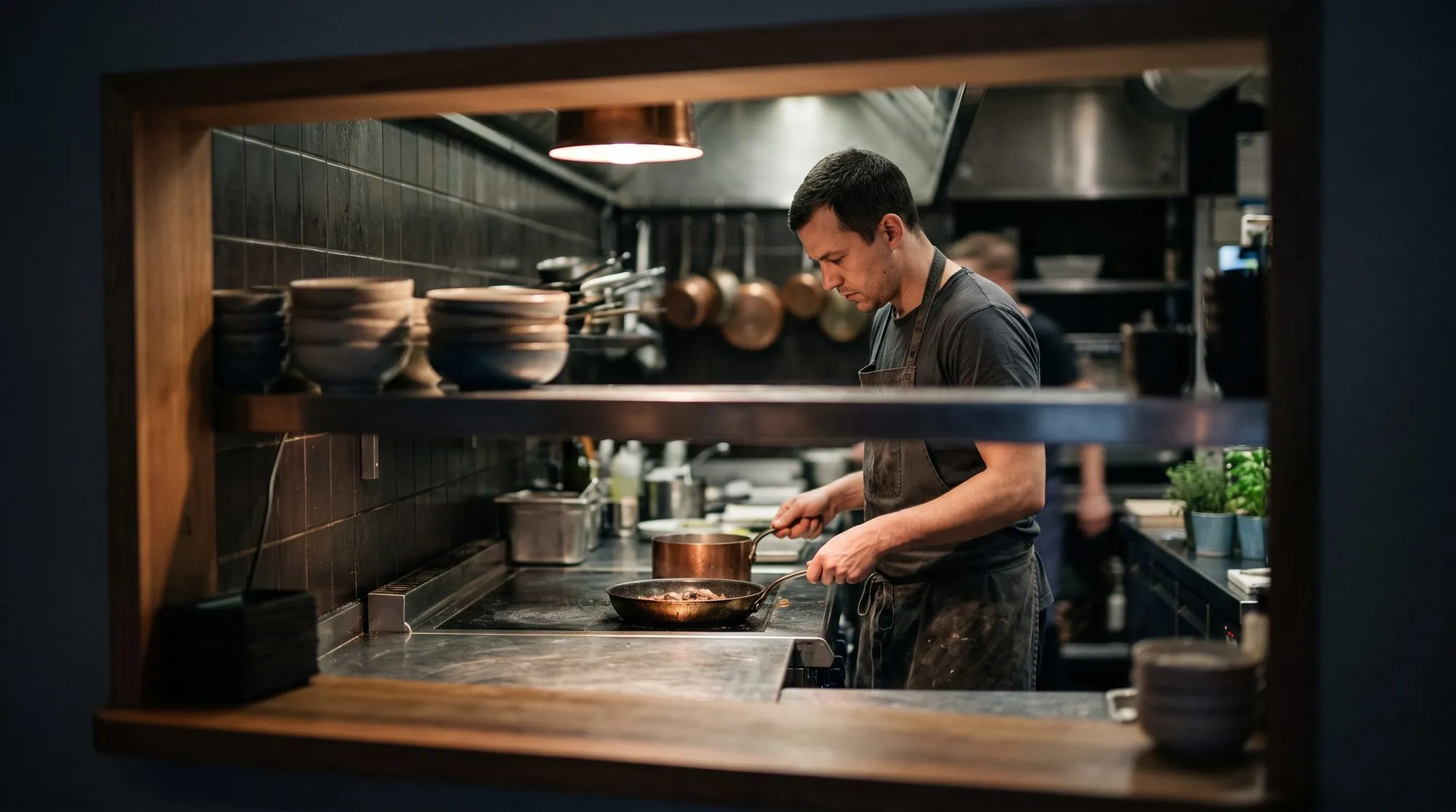 A cook at the stove seen through a bistro service window, copper pans on the shelf above