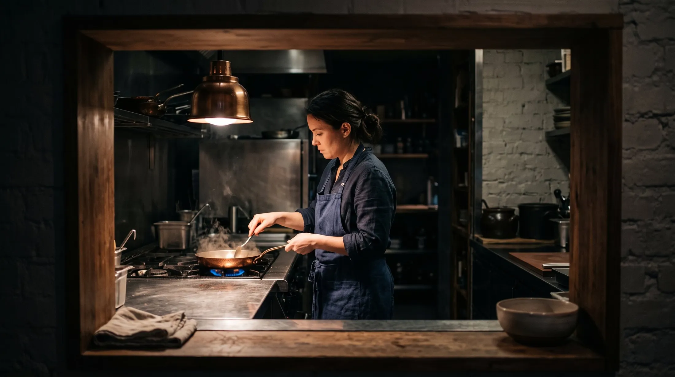 A cook at a stove in a dark kitchen, one warm pool of light